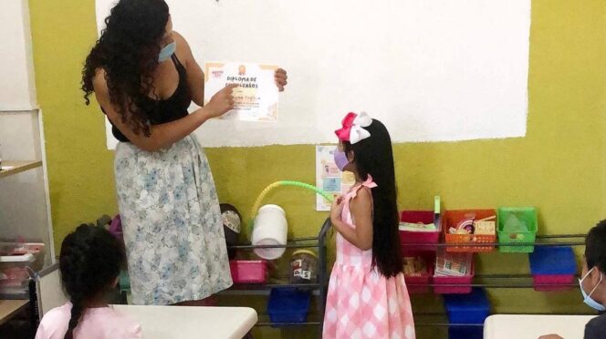 Teacher presents a diploma to a young girl in a colorful classroom while peers watch, both wearing masks.