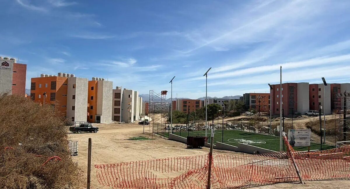 Construction site with orange and beige apartment buildings under a blue sky; a small fenced artificial-turf soccer field on the right.