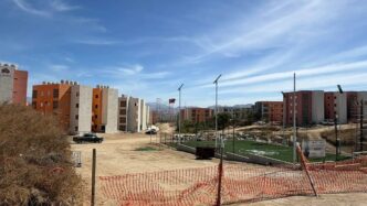 Construction site with orange and beige apartment buildings under a blue sky; a small fenced artificial-turf soccer field on the right.