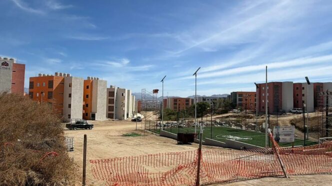 Construction site with orange and beige apartment buildings under a blue sky; a small fenced artificial-turf soccer field on the right.