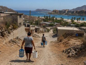 Two women walking down a rocky dirt road carrying large water jugs toward a coastal town and beach. Proximity to ocean and hotels hints at a seaside community effort to fetch water.