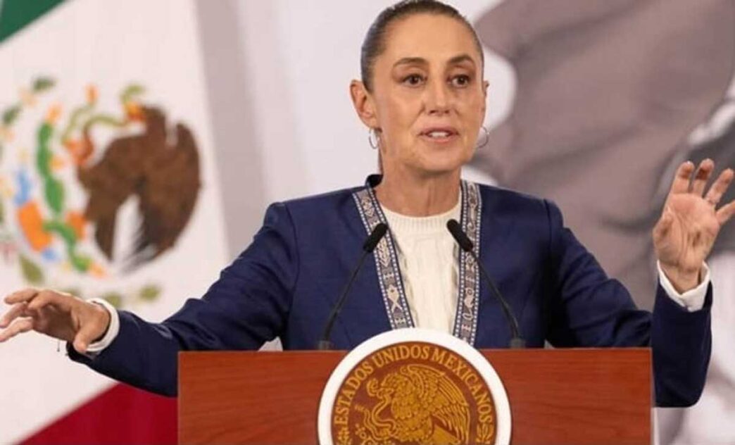 Woman speaking at a wooden podium featuring the Mexican seal, gesturing with both hands during a formal address behind microphones.