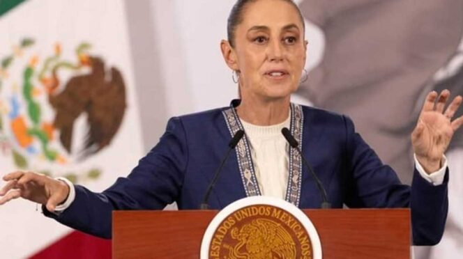 Woman speaking at a wooden podium featuring the Mexican seal, gesturing with both hands during a formal address behind microphones.