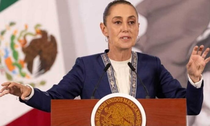 Woman speaking at a wooden podium featuring the Mexican seal, gesturing with both hands during a formal address behind microphones.