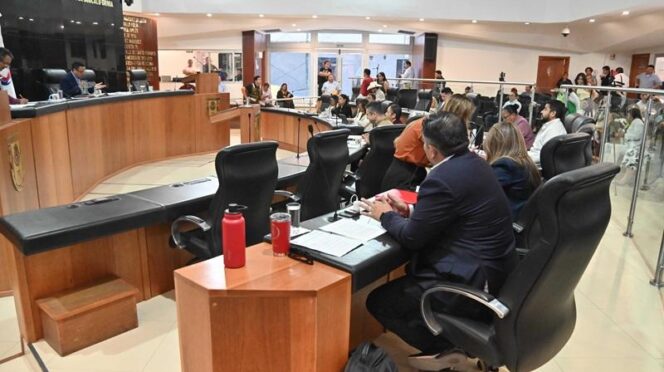 City council chamber with officials seated around a curved desk, microphones and documents visible.