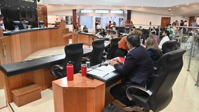 City council chamber with officials seated around a curved desk, microphones and documents visible.
