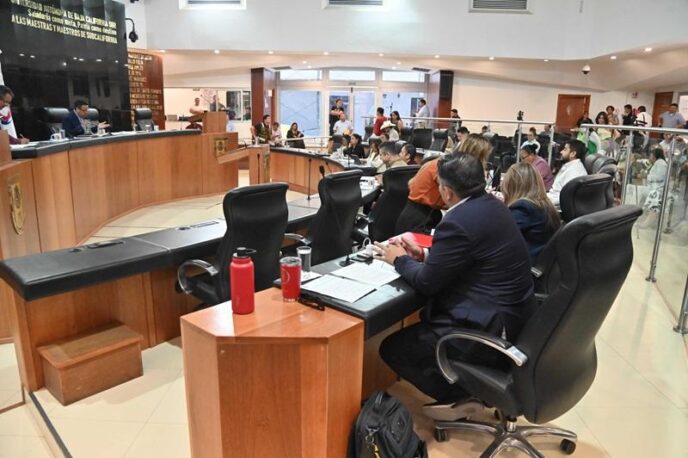 City council chamber with officials seated around a curved desk, microphones and documents visible.