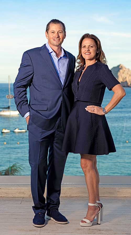 dennis-renee-couple-hair3-1 - Gringo Gazette Well-dressed couple posing on a balcony by the sea; man in a blue suit and woman in a navy dress, smiling at the camera.