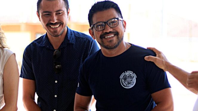 Two men smiling at the camera, one wearing a navy dotted shirt with sunglasses tucked in his chest and the other in a dark t-shirt with a circular emblem on the chest, bright background behind them.