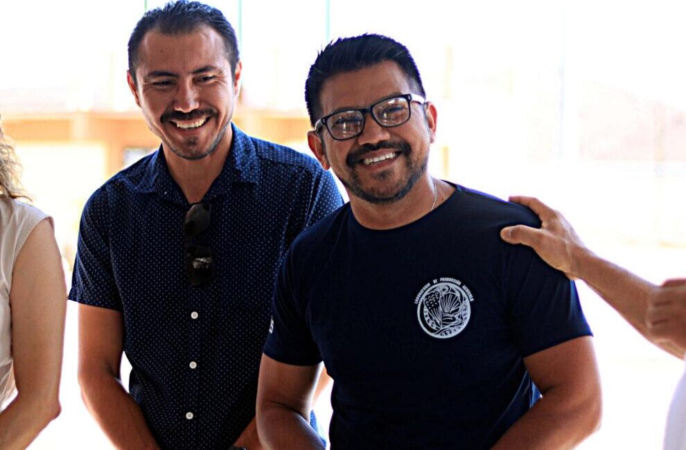 Two men smiling at the camera, one wearing a navy dotted shirt with sunglasses tucked in his chest and the other in a dark t-shirt with a circular emblem on the chest, bright background behind them.