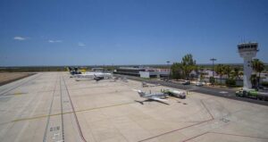 Wide view of a small airport tarmac with a plane at a gate, fueling trucks nearby, and a control tower in the distance under a clear blue sky.