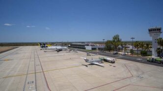 Wide view of a small airport tarmac with a plane at a gate, fueling trucks nearby, and a control tower in the distance under a clear blue sky.