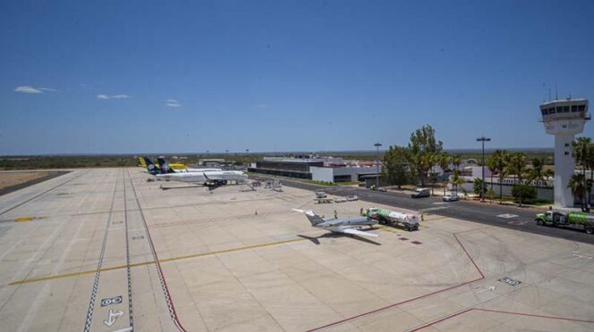 Wide view of a small airport tarmac with a plane at a gate, fueling trucks nearby, and a control tower in the distance under a clear blue sky.
