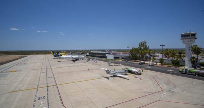Wide view of a small airport tarmac with a plane at a gate, fueling trucks nearby, and a control tower in the distance under a clear blue sky.