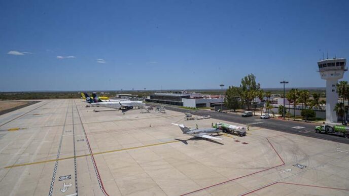 Wide view of a small airport tarmac with a plane at a gate, fueling trucks nearby, and a control tower in the distance under a clear blue sky.