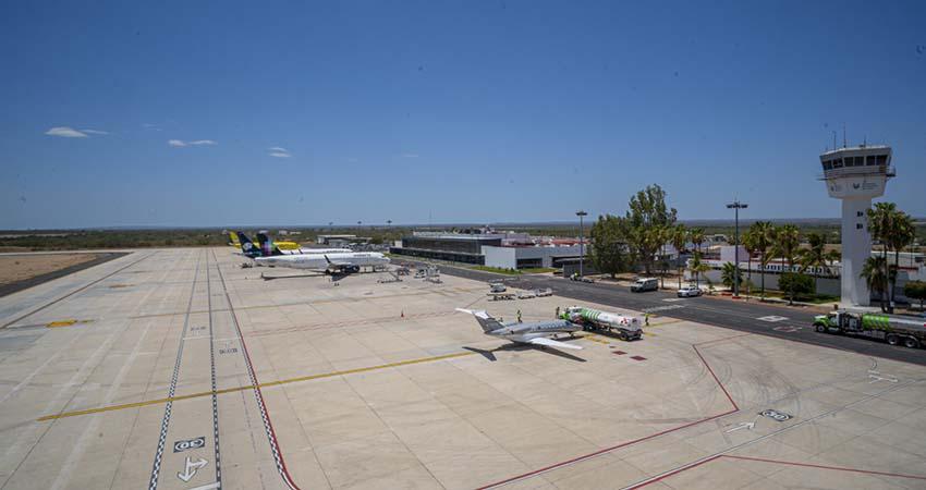 Wide view of a small airport tarmac with a plane at a gate, fueling trucks nearby, and a control tower in the distance under a clear blue sky.