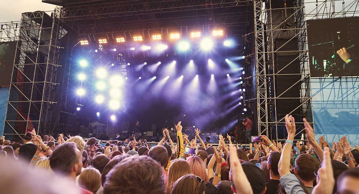 Large outdoor concert stage with bright lights and a cheering crowd raising their hands in front of it.