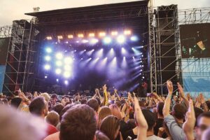 Large outdoor concert stage with bright lights and a cheering crowd raising their hands in front of it.
