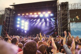 Large outdoor concert stage with bright lights and a cheering crowd raising their hands in front of it.