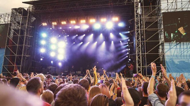 Large outdoor concert stage with bright lights and a cheering crowd raising their hands in front of it.