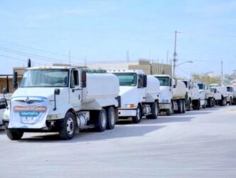 Line of white delivery trucks driving in a convoy down a city street under a clear sky.