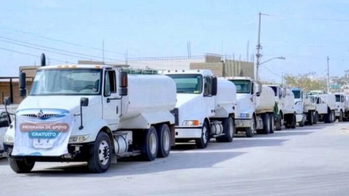 Line of white delivery trucks driving in a convoy down a city street under a clear sky.