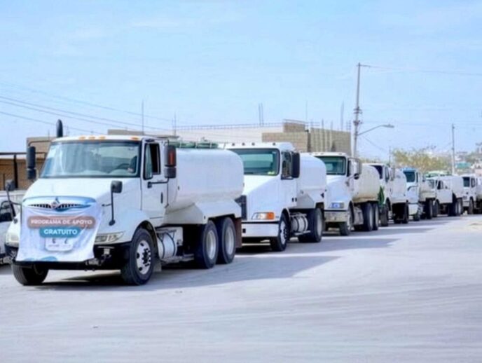 Line of white delivery trucks driving in a convoy down a city street under a clear sky.