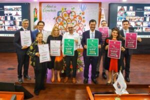 Ten people in business attire stand on a stage, each holding a certificate of achievement in different colors, with a decorative mural behind.