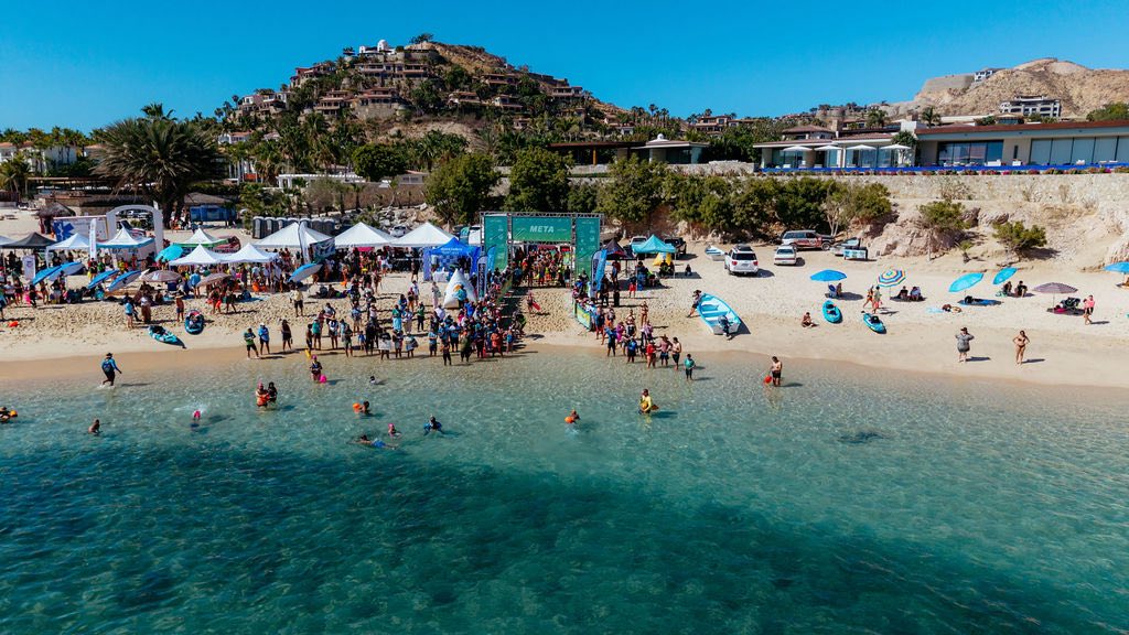 Crowded beach event with white tents, a green META arch, and swimmers in clear turquoise water near the shore.