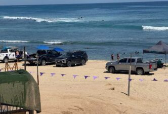 Cars and trucks parked on a sandy beach beside the ocean, with purple pennant flags in the foreground.