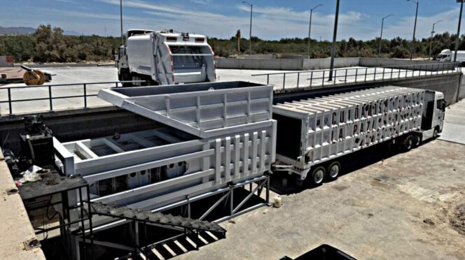 Industrial loading yard with a white refuse truck and a long white trailer beside a large metal container structure at a dock-like site.