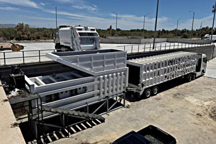 Industrial loading yard with a white refuse truck and a long white trailer beside a large metal container structure at a dock-like site.