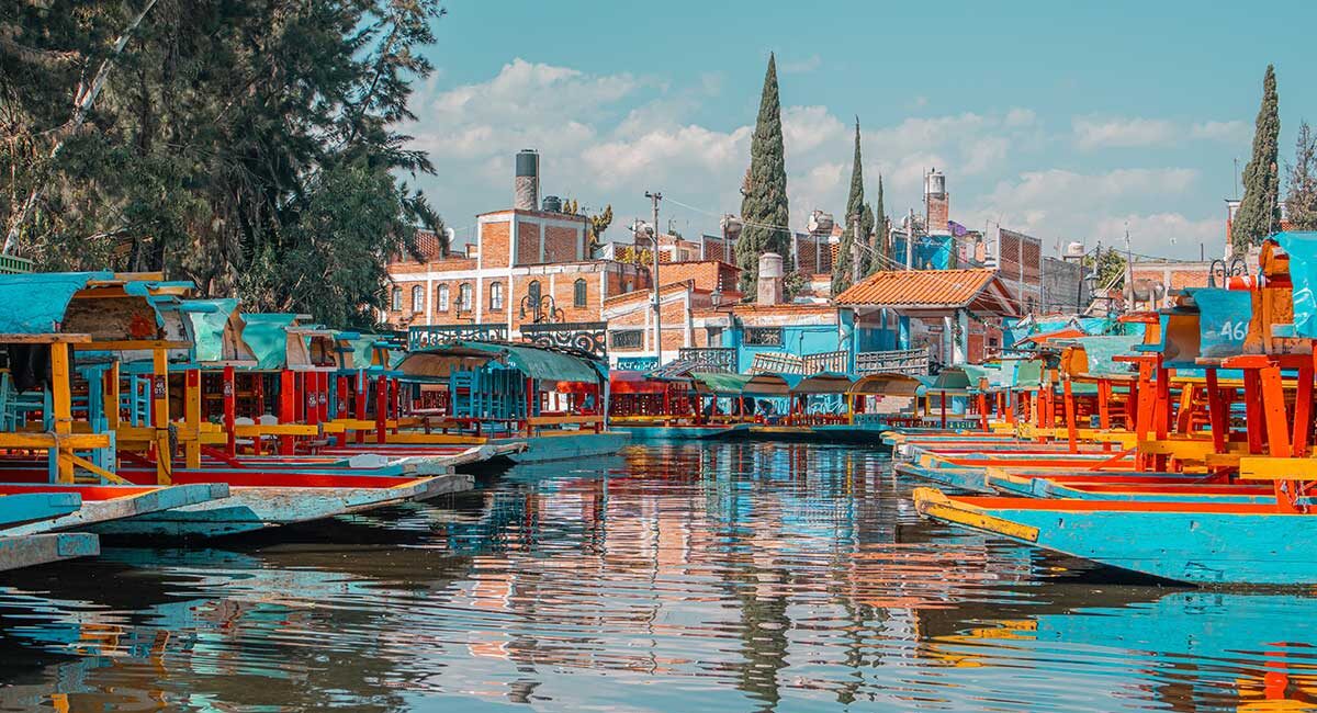 Colorful trajinera boats docked along a canal with reflections in the water and red-brick buildings in the background.