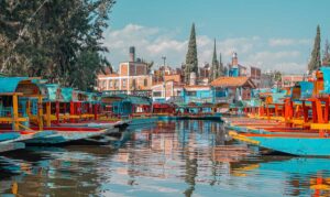 Colorful trajinera boats docked along a canal with reflections in the water and red-brick buildings in the background.