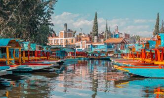 Colorful trajinera boats docked along a canal with reflections in the water and red-brick buildings in the background.