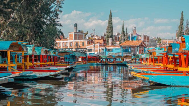 Colorful trajinera boats docked along a canal with reflections in the water and red-brick buildings in the background.