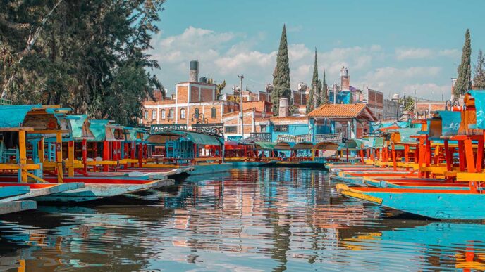 Colorful trajinera boats docked along a canal with reflections in the water and red-brick buildings in the background.