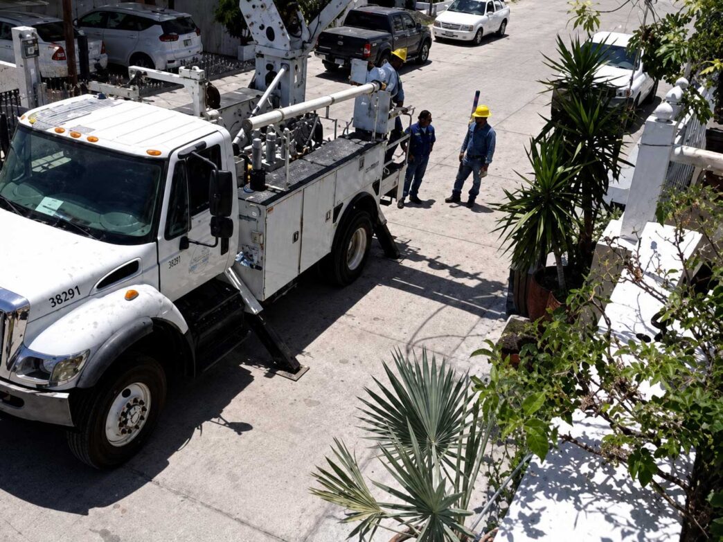 White utility truck with a tall bucket lift parked on a sunny street as two workers in yellow hard hats discuss nearby.