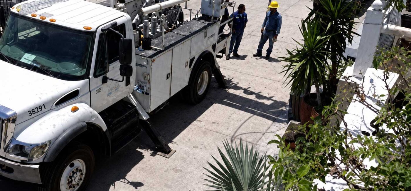 White utility truck with a tall bucket lift parked on a sunny street as two workers in yellow hard hats discuss nearby.