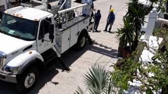 White utility truck with a tall bucket lift parked on a sunny street as two workers in yellow hard hats discuss nearby.