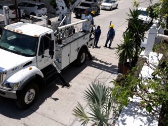 White utility truck with a tall bucket lift parked on a sunny street as two workers in yellow hard hats discuss nearby.
