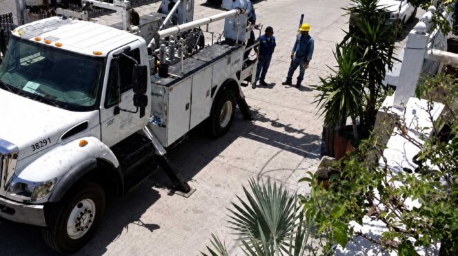White utility truck with a tall bucket lift parked on a sunny street as two workers in yellow hard hats discuss nearby.