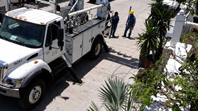 White utility truck with a tall bucket lift parked on a sunny street as two workers in yellow hard hats discuss nearby.