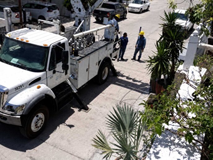White utility truck with a tall bucket lift parked on a sunny street as two workers in yellow hard hats discuss nearby.
