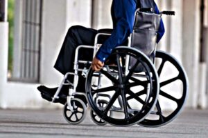 Person in a wheelchair being pushed along a city sidewalk, urban background visible in the distance.