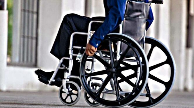 Person in a wheelchair being pushed along a city sidewalk, urban background visible in the distance.