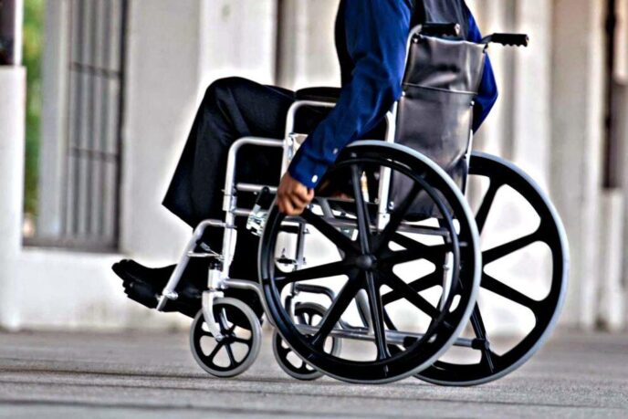 Person in a wheelchair being pushed along a city sidewalk, urban background visible in the distance.