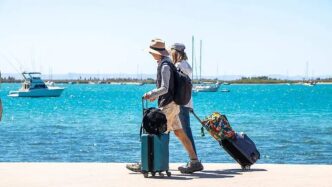 Couple walking along a marina with rolling suitcases and backpacks by bright blue water and boats in the background.