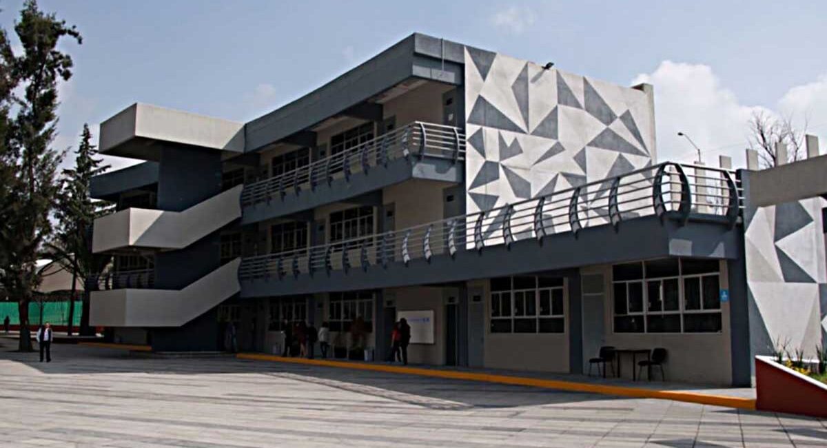 Modern grey building with external zigzag staircases and a geometric triangular mural on the side; people near the entrance on a paved plaza.