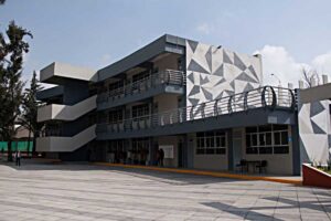 Modern grey building with external zigzag staircases and a geometric triangular mural on the side; people near the entrance on a paved plaza.